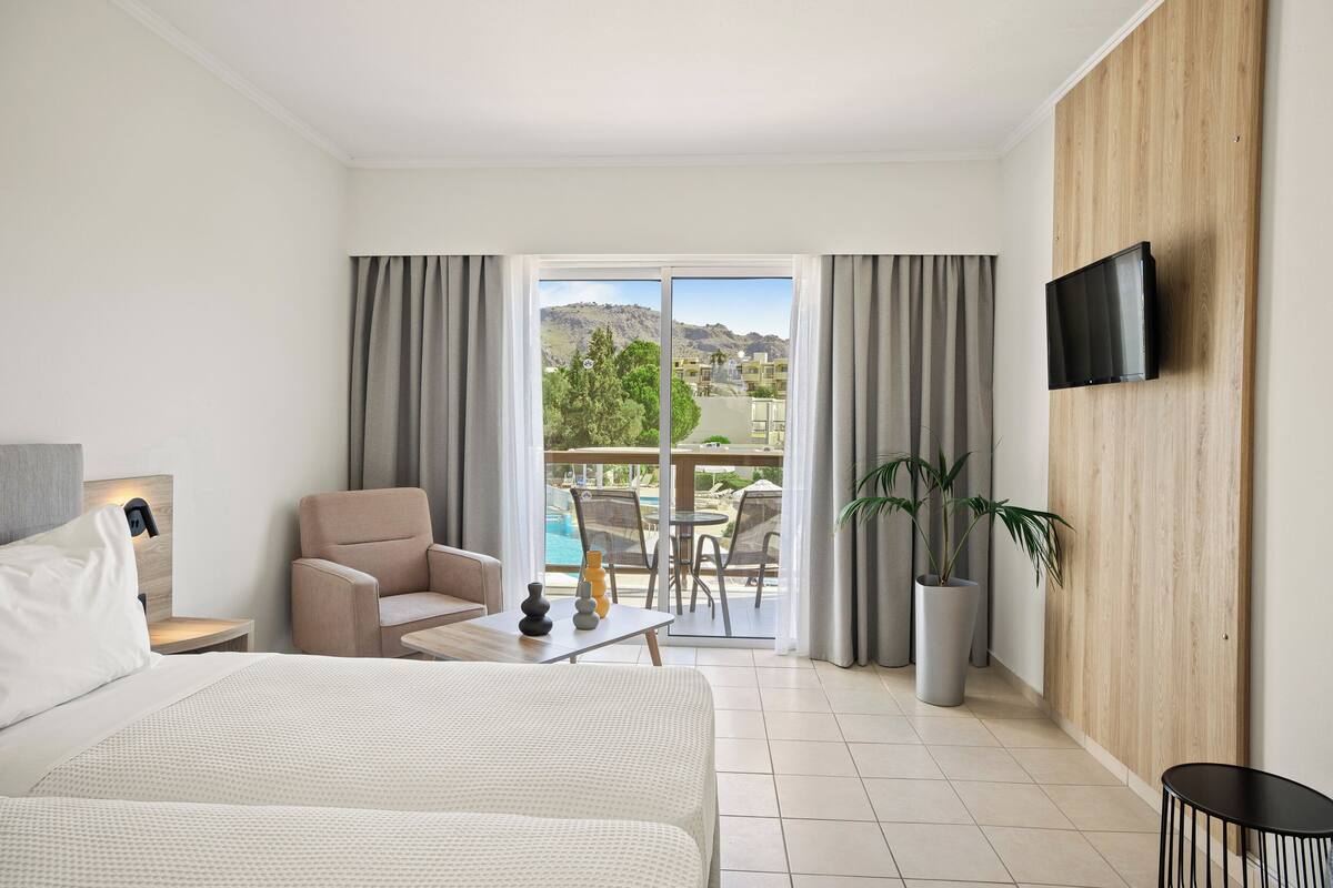 Une chambre d’hôtel avec un lit, un canapé, un téléviseur fixé au mur, un balcon donnant sur une piscine et une vue sur les montagnes.