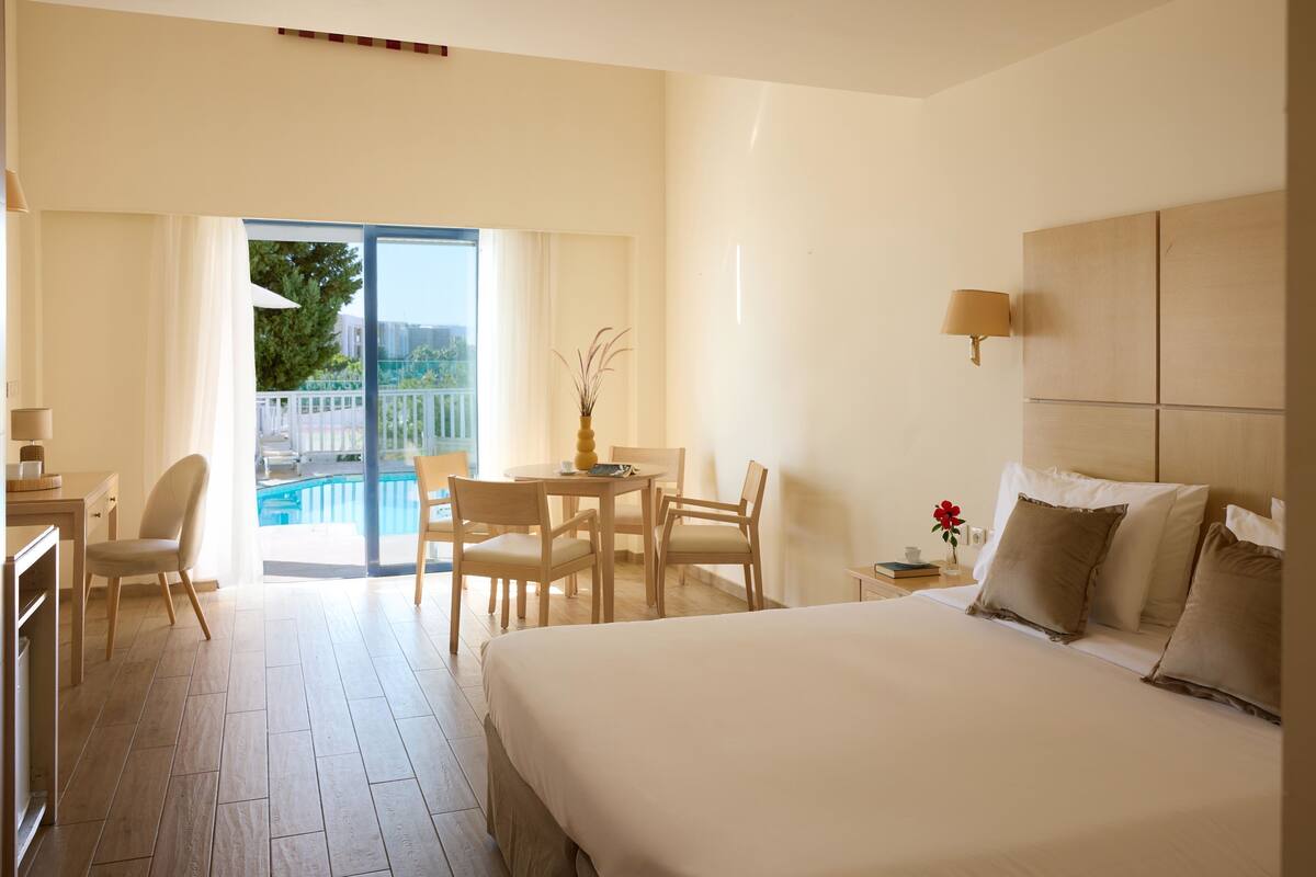 Une chambre d’hôtel avec un grand lit, un bureau, une chaise, une petite table et une vue sur une piscine et un espace verdoyant.