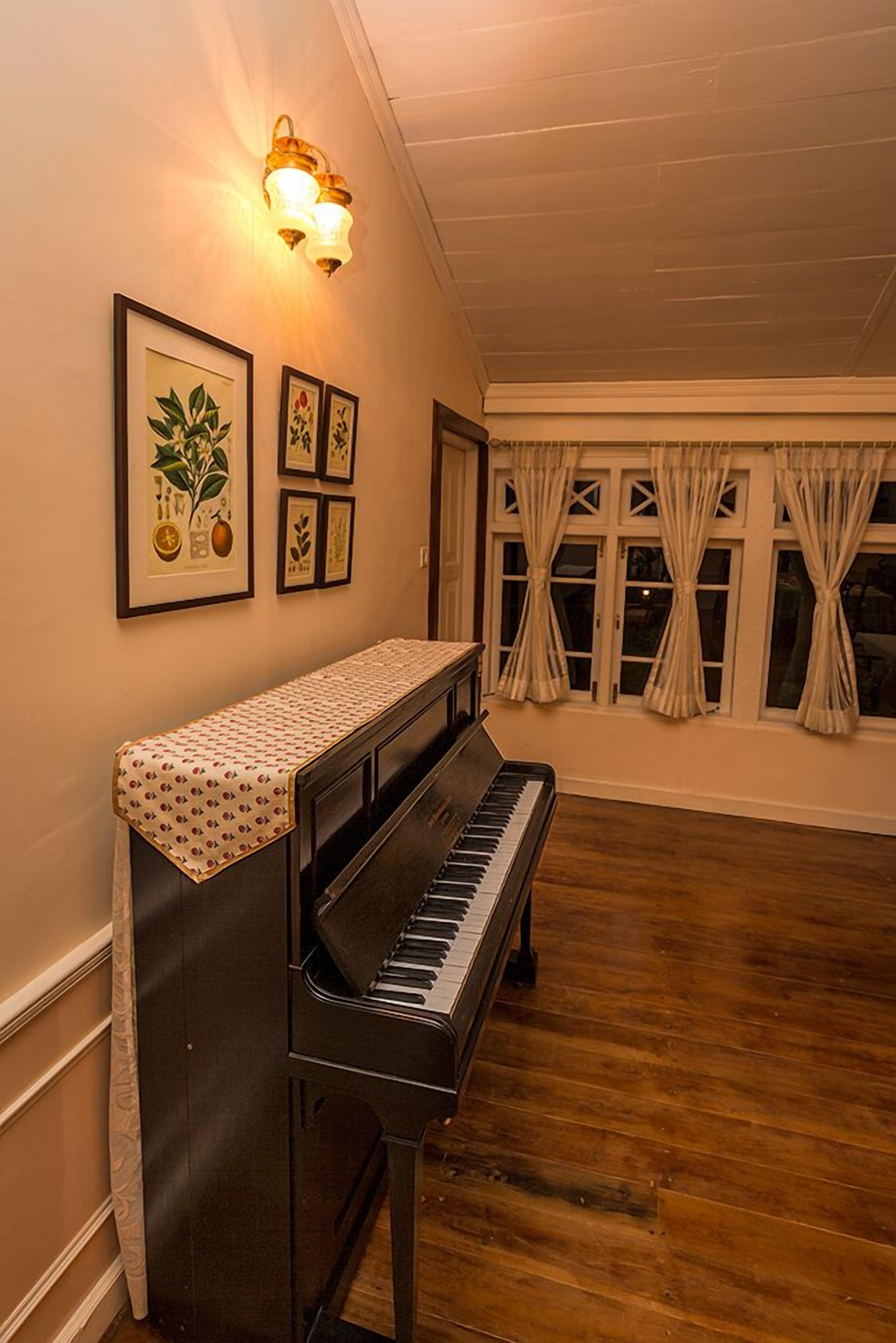 A piano with a patterned cloth cover, a wall-mounted light fixture, and framed botanical illustrations on the wall.