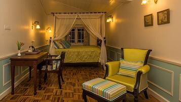Una habitación con piso de parquet de madera, cama con dosel, sillón amarillo, una mesita con un florero e iluminación en la pared.