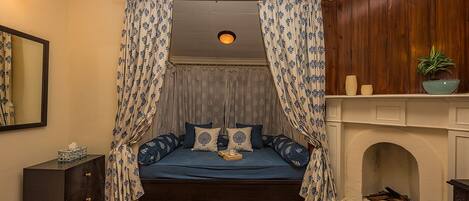 A bedroom with a bed, curtains, a dresser, a mirror, a fireplace, and a ceiling light.