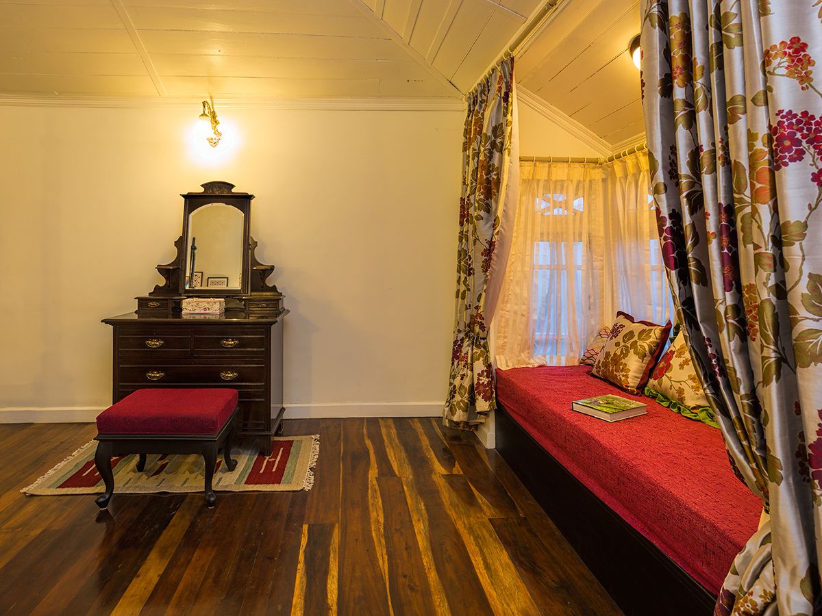 Une chambre avec un plancher en bois, un lit rouge, une commode avec un miroir et des rideaux à motifs floraux.