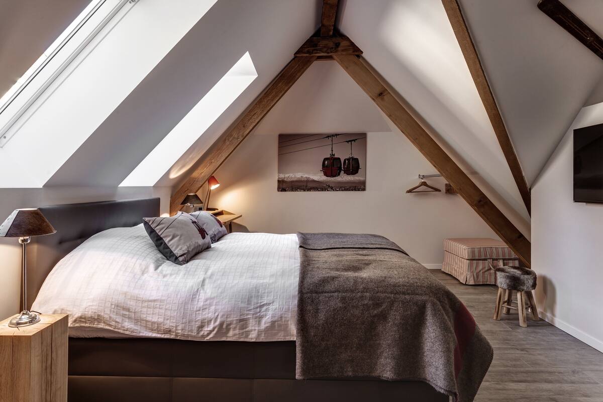 A bedroom with a sloped ceiling, a bed with a headboard, a bedside table with a lamp, a hanging coat rack, a small stool, and a framed picture on the wall.