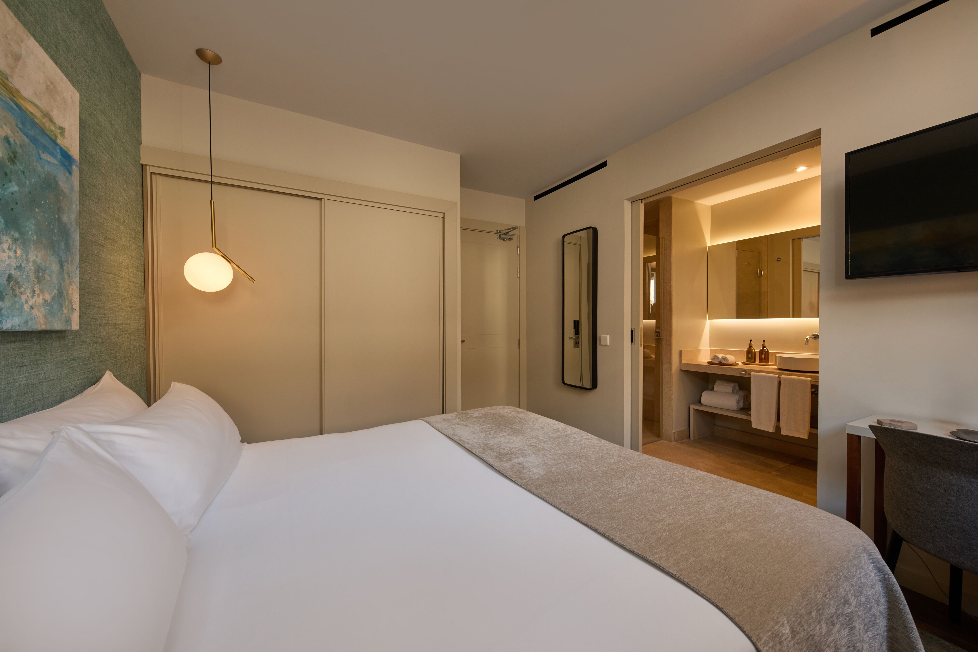 Ein modernes Hotelzimmer mit einem ordentlich bezogenen Bett, einem Badezimmer mit Spiegel und Waschbecken sowie einem an der Wand befestigten Fernseher.