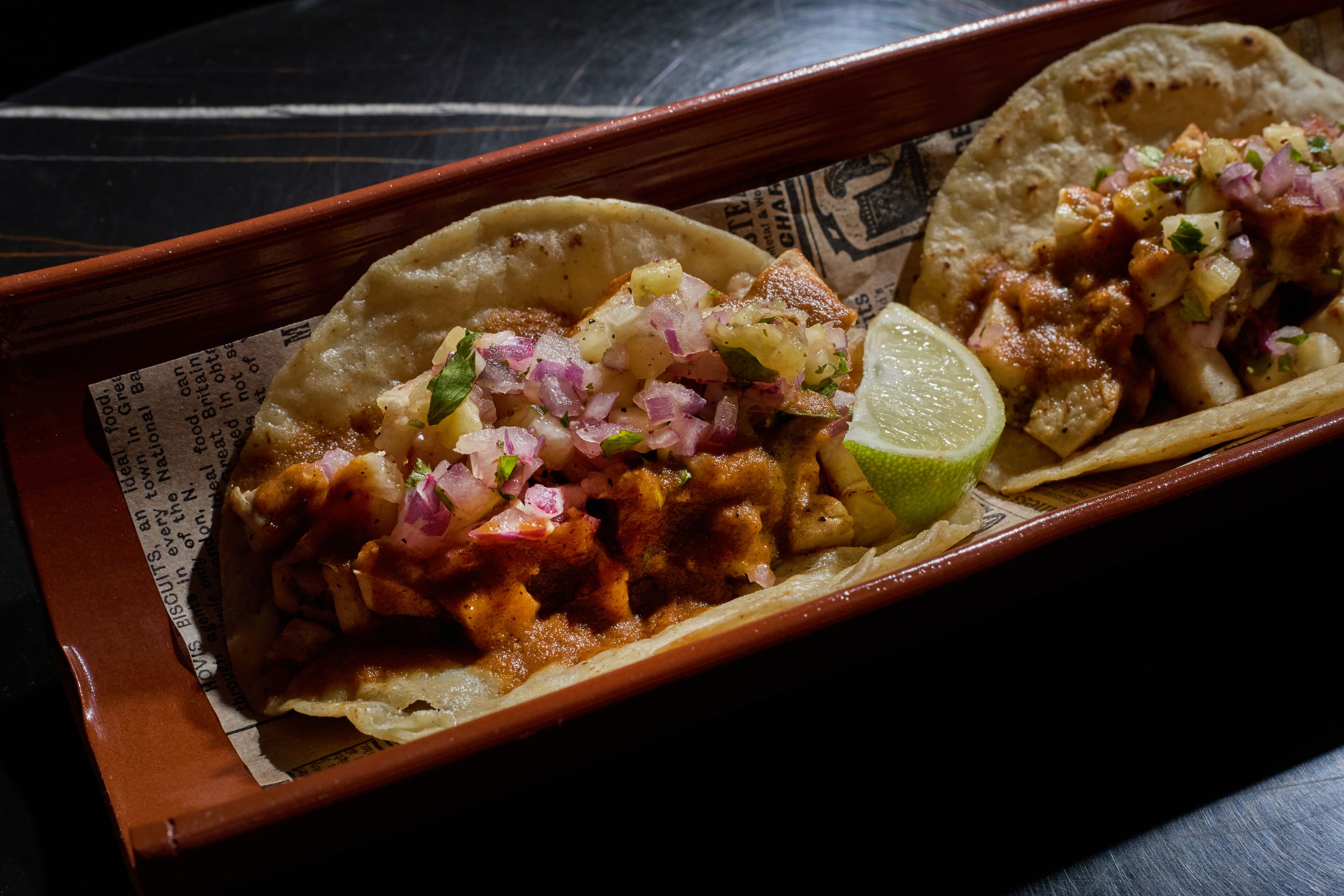 Una bandeja con dos tacos, uno con una cuña de lima, y un periódico debajo.