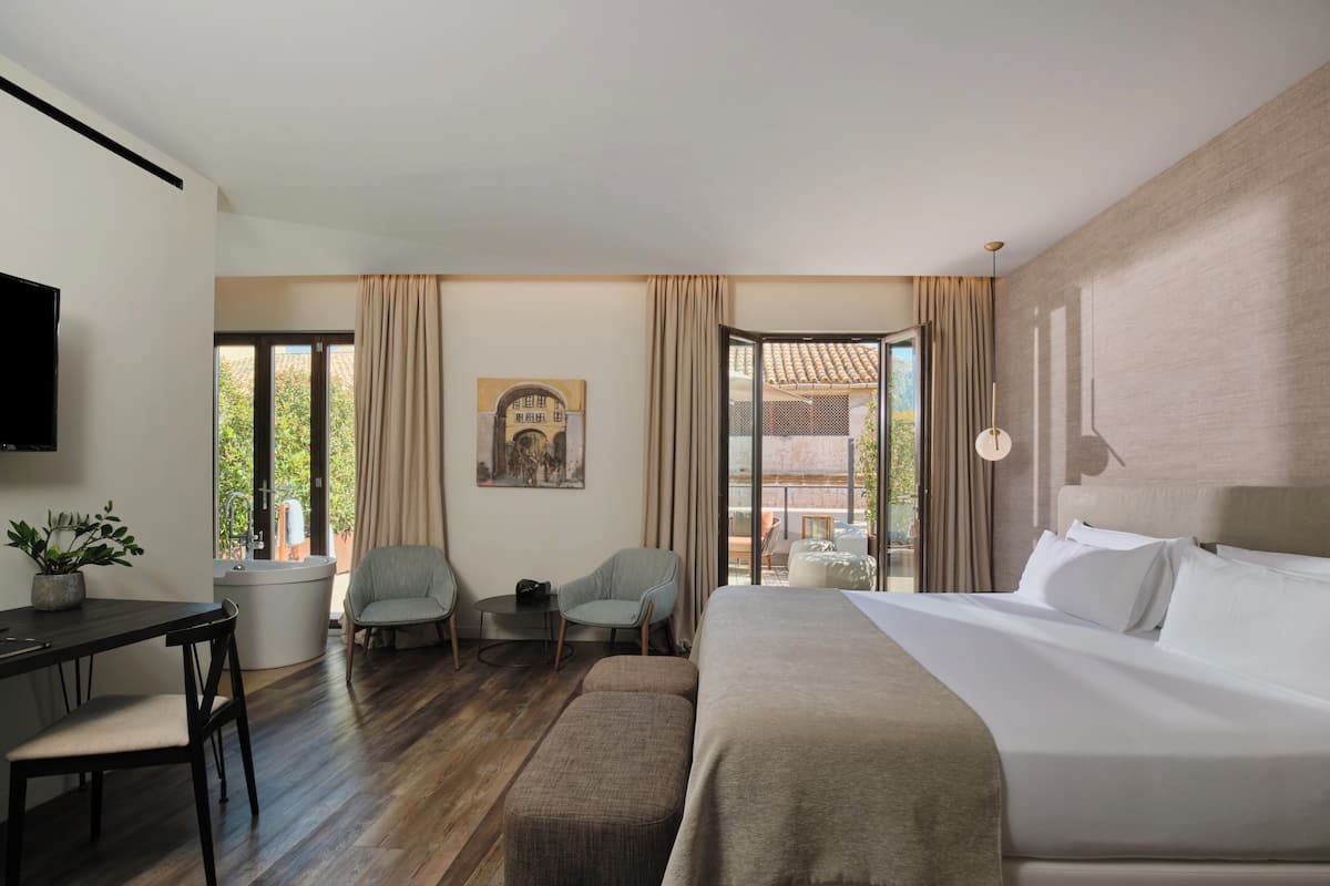 Una habitación de hotel moderna con una cama grande, un escritorio con una silla, una mesita pequeña y un sofá. Hay un televisor montado en la pared, una ventana con cortinas y un cuadro en la pared.