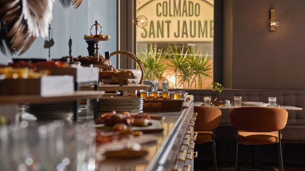 Un restaurante con bufé, zona de estar y un ventanal con el nombre "Colmado Sant Jaume".