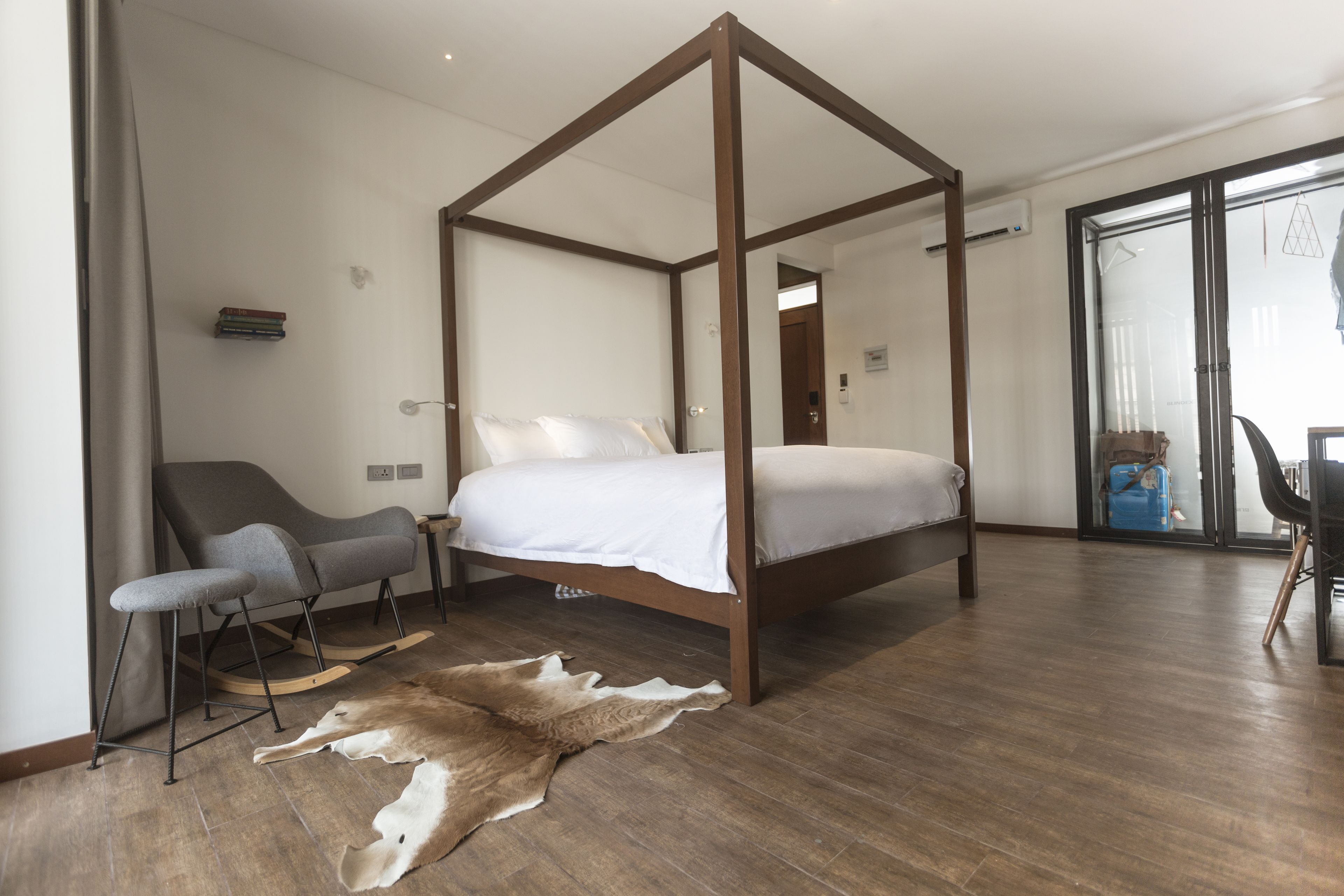 A modern bedroom with a four-poster bed, a grey armchair, a small bench, and a fur rug on the wooden floor.