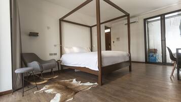 A modern bedroom with a four-poster bed, a grey armchair, a small bench, and a fur rug on the wooden floor.
