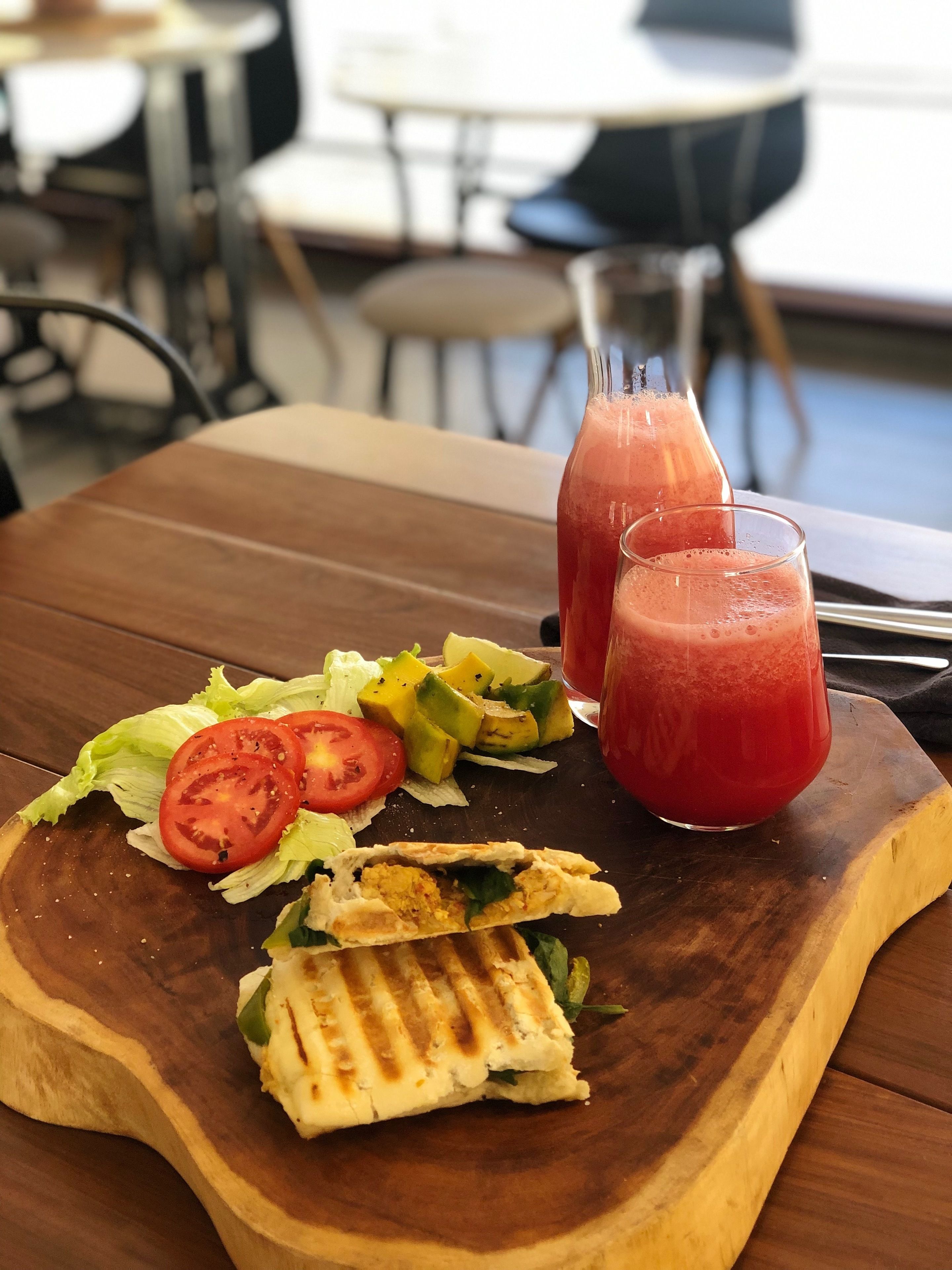 Una tabla de madera con un sándwich a la plancha, verduras frescas y dos vasos de jugo de mora.