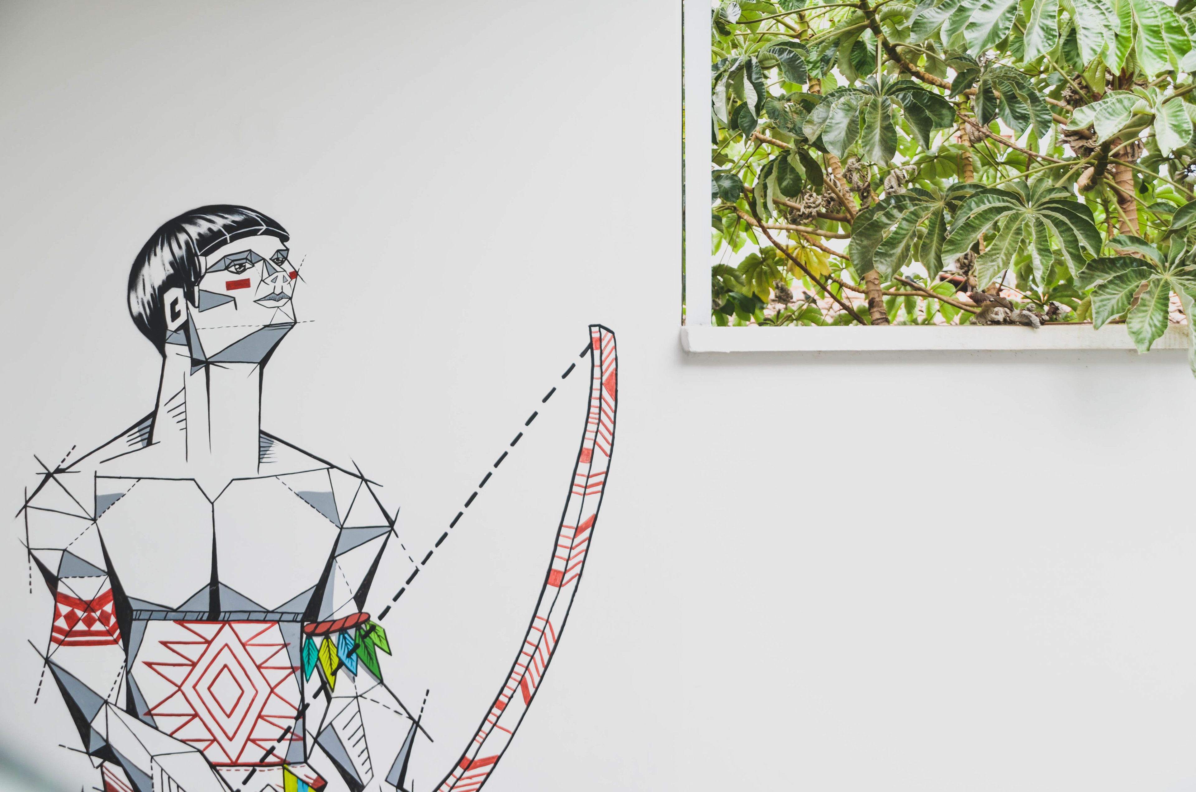 Una figura humana geométrica con un diseño rojo y blanco, una planta de hojas verdes y una pared blanca.