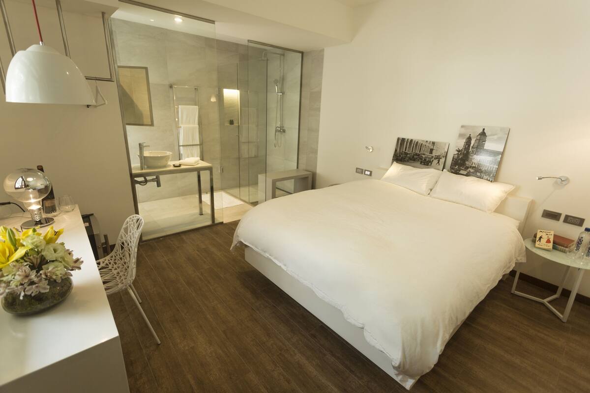 A modern hotel room with a large bed, a glass-enclosed shower, a desk with a lamp, and a small table with a book and a bottle.