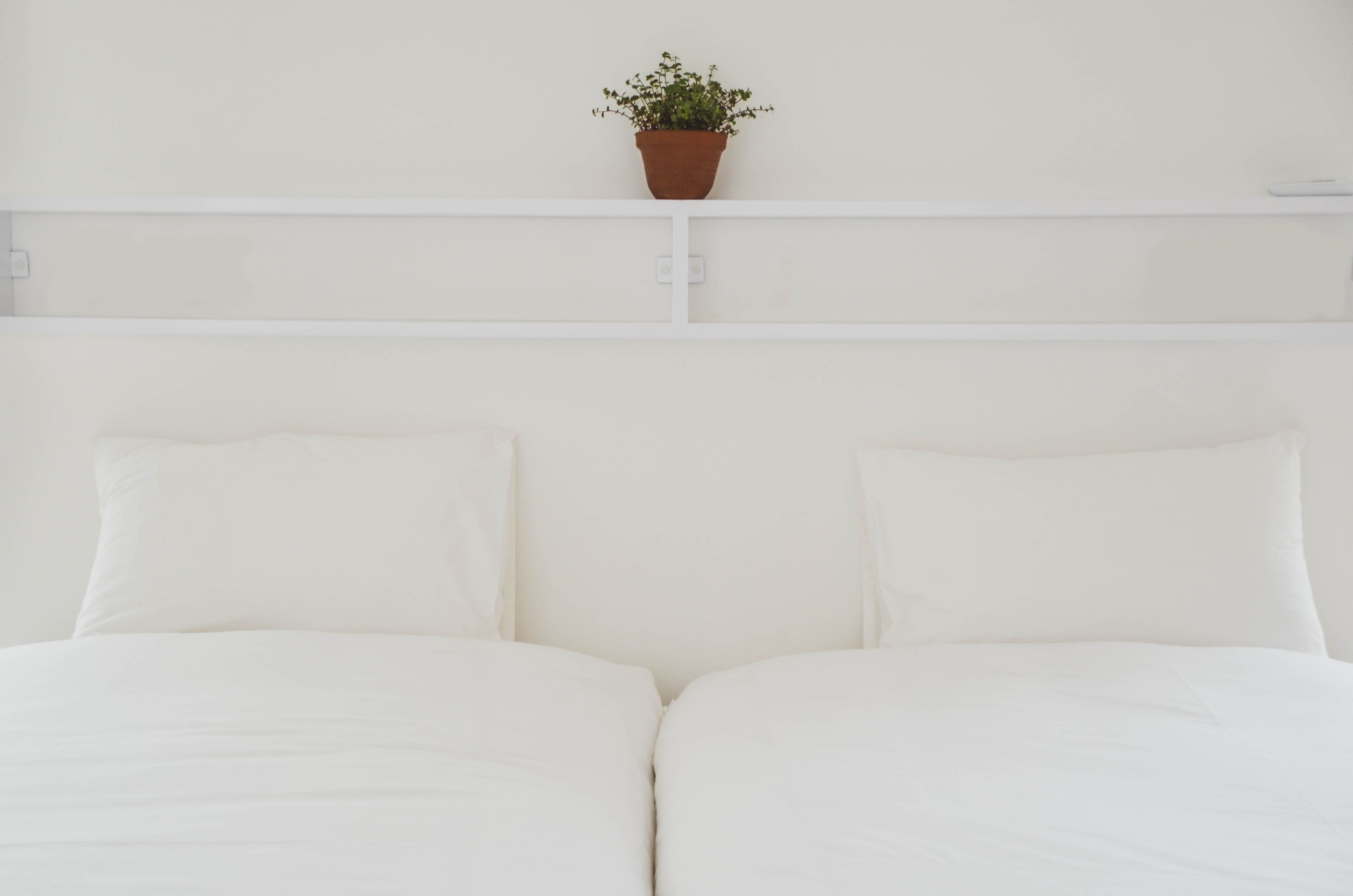 Una cama con ropa de cama blanca y una planta en maceta sobre un cabecero blanco.