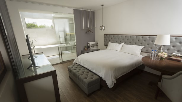 A modern hotel room with a large bed, a bedside table with a lamp, a small bench, and a glass-enclosed bathroom. - Cosmopolitano Hotel Boutique (Santa Cruz)