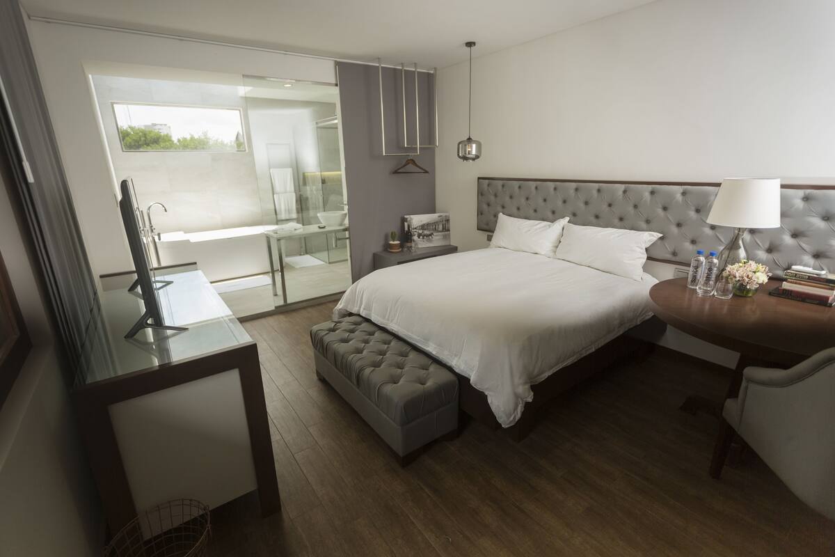 A modern hotel room with a large bed, a bedside table with a lamp, a small bench, and a glass-enclosed bathroom.