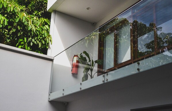 Un edificio moderno con balcón de vidrio, un extintor y abundante follaje verde.