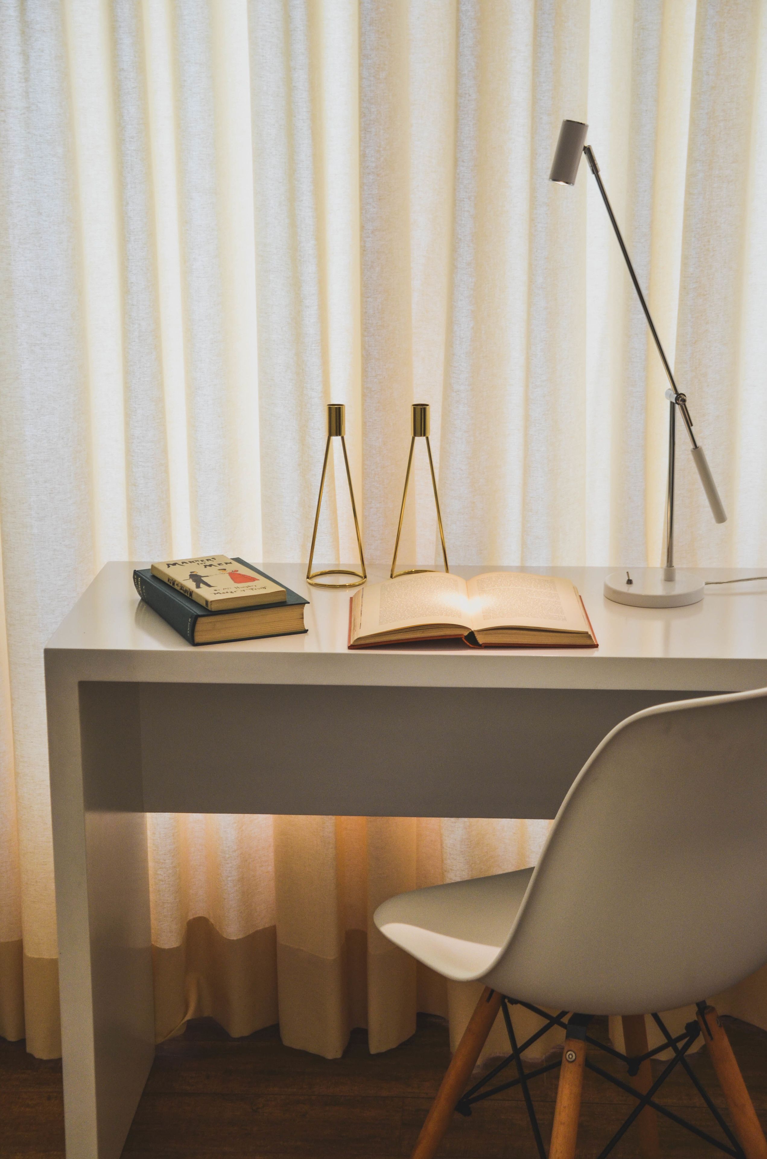 A desk with a chair, an open book, two lamps, and a pair of books on the desk.