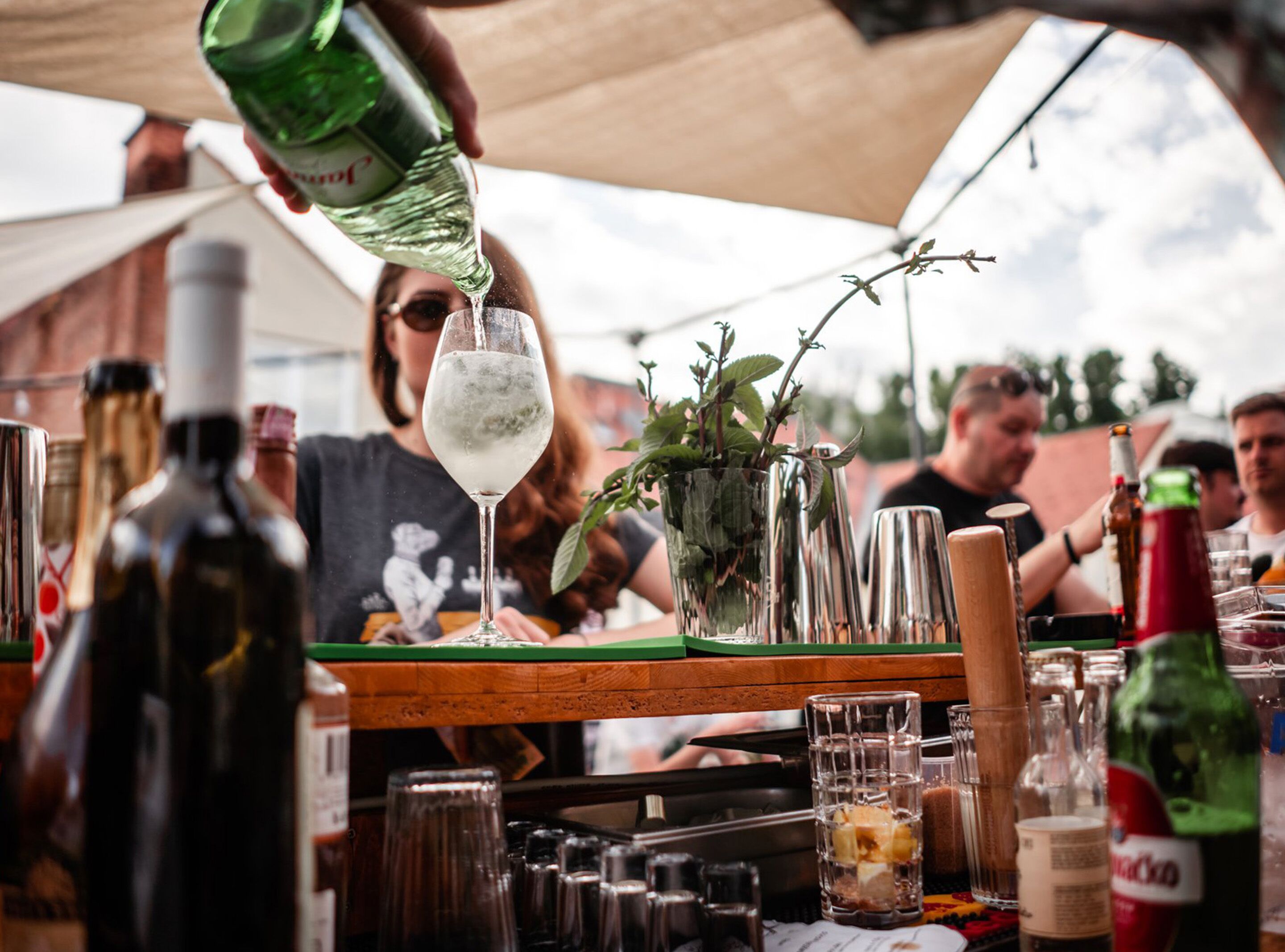 Un bar con diversas botellas y vasos, una mesa de madera y una persona sirviendo una bebida.