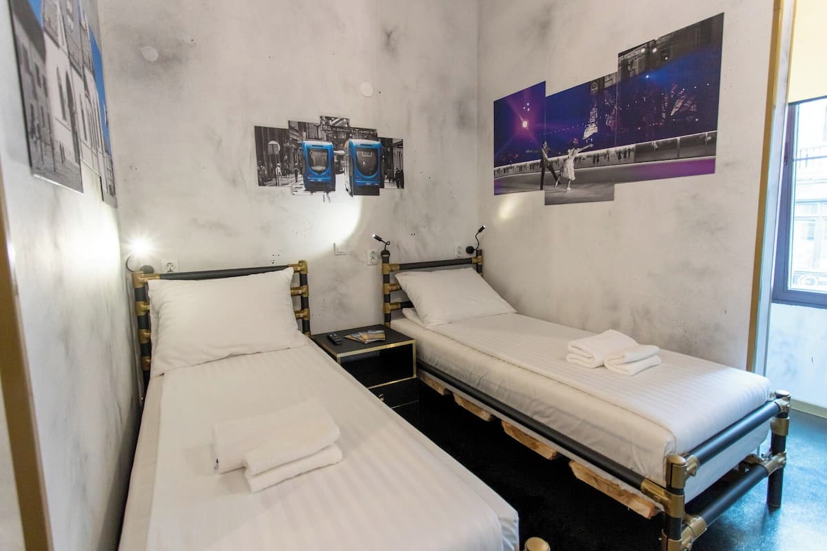 Ein Zimmer mit zwei Betten, jeweils mit Holzrahmen und weißer Bettwäsche. Zwei Nachttische mit Lampen und ein kleiner Tisch zwischen den Betten. Die Wände sind schlicht gehalten, mit einigen Postern und einem großen Fenster auf der rechten Seite.