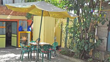 Un área de estar al aire libre con una mesa y sillas bajo un paraguas amarillo, rodeada de follaje y un muro de ladrillo.