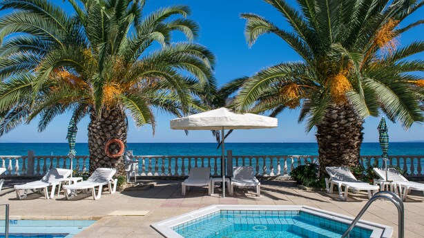 A poolside area with lounge chairs, a pool, and palm trees. - Paradise Beach Hotel (Zakynthos)