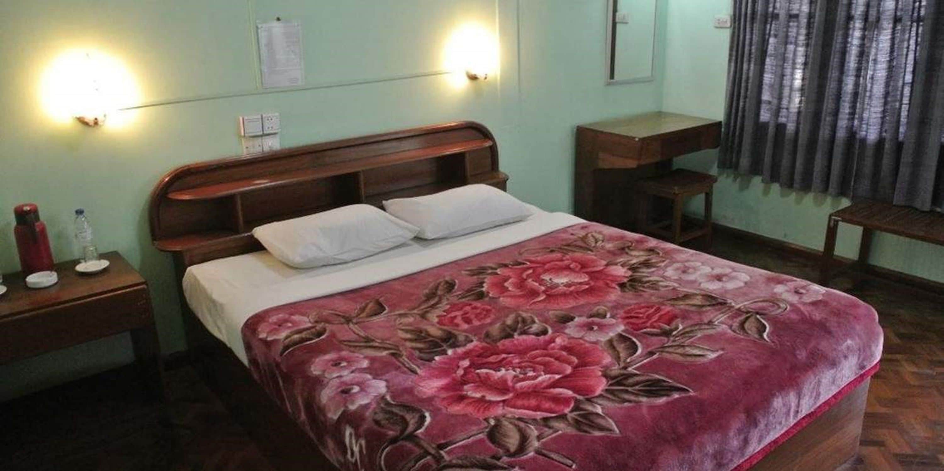 A bedroom with a bed, two bedside tables, a nightstand, a wooden headboard, a floral bedspread, and a window with curtains.