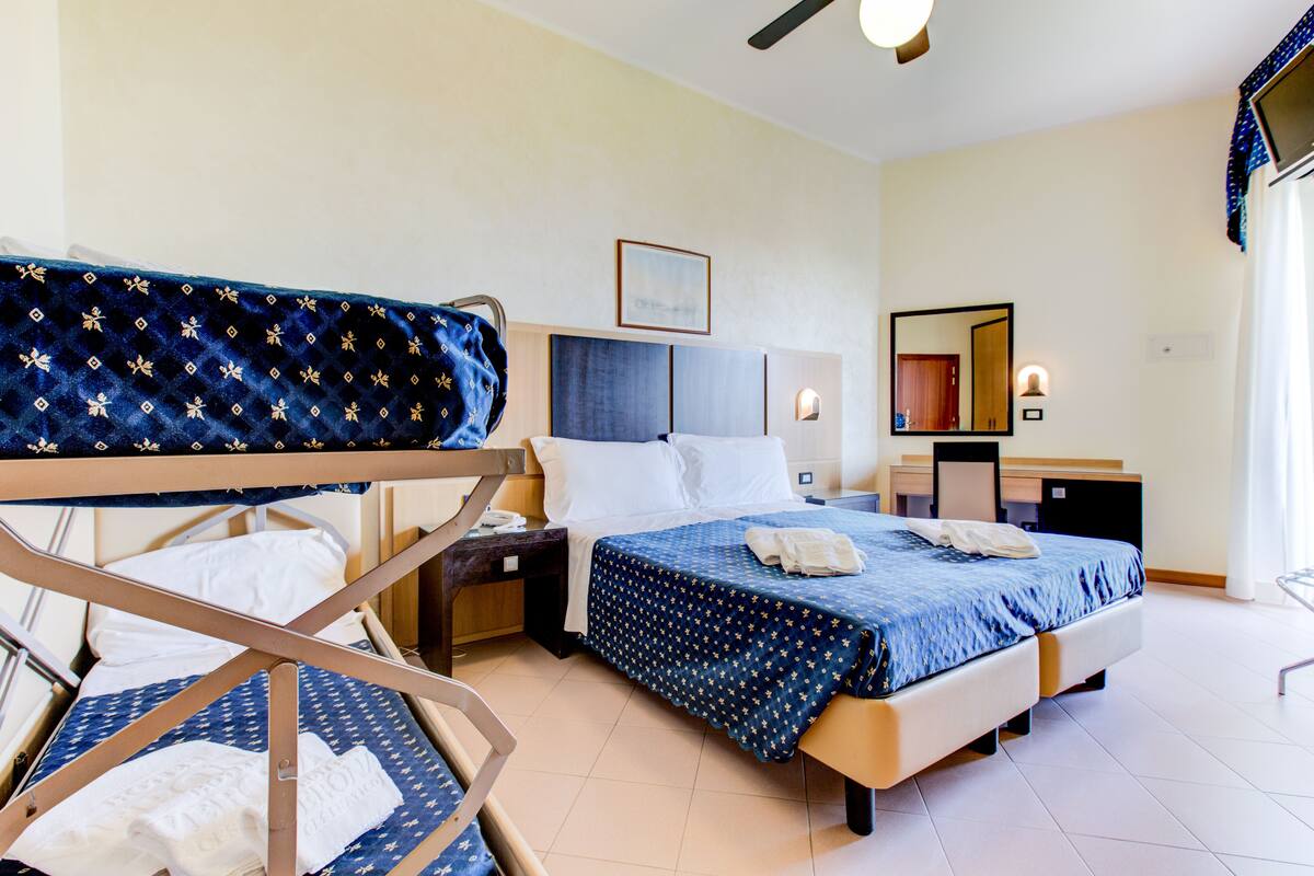 A hotel room with a bunk bed, a single bed, a desk with a chair, a bedside table, and a mirror.
