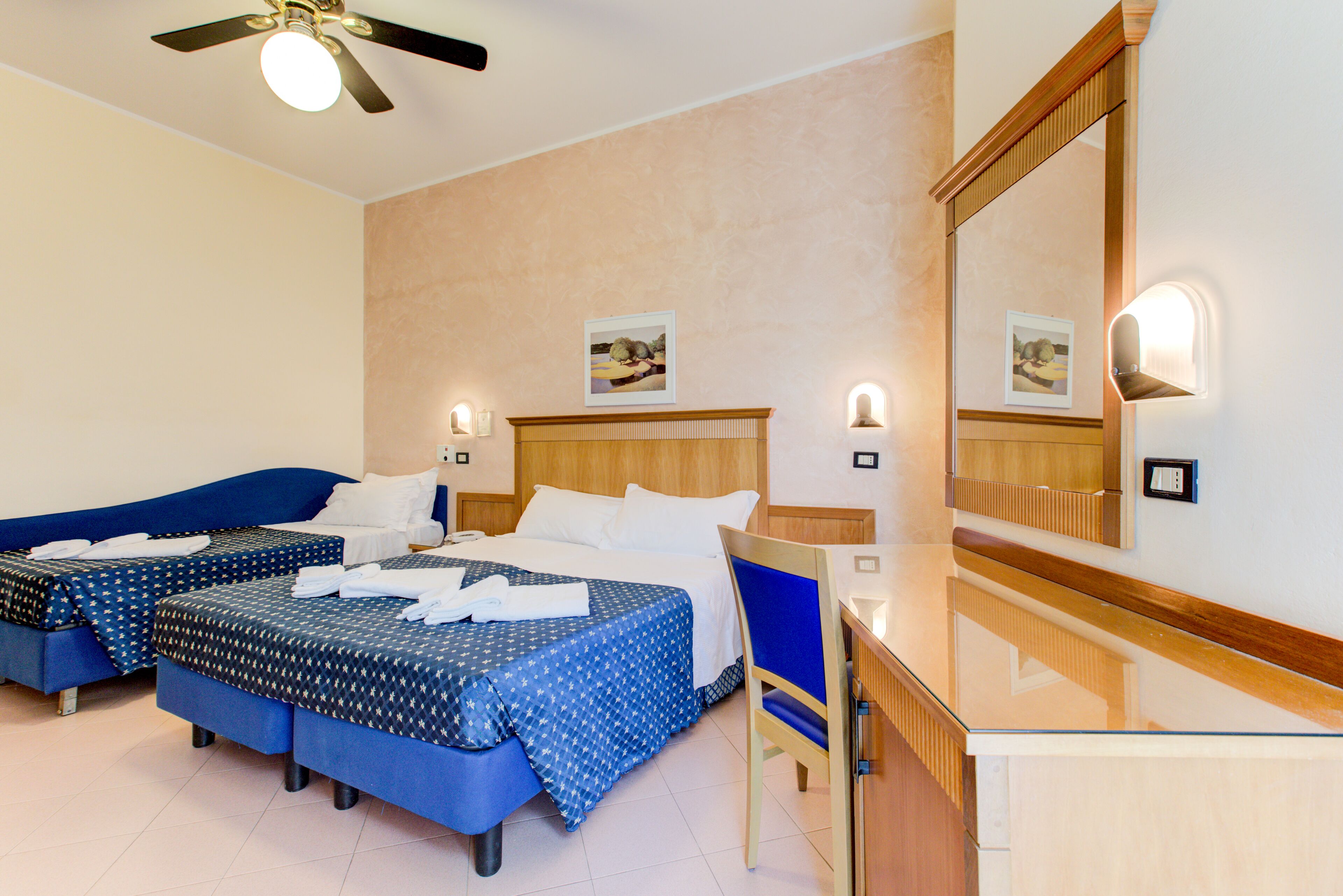 A hotel room with two beds, a desk, a chair, a ceiling fan, and wall-mounted lights.