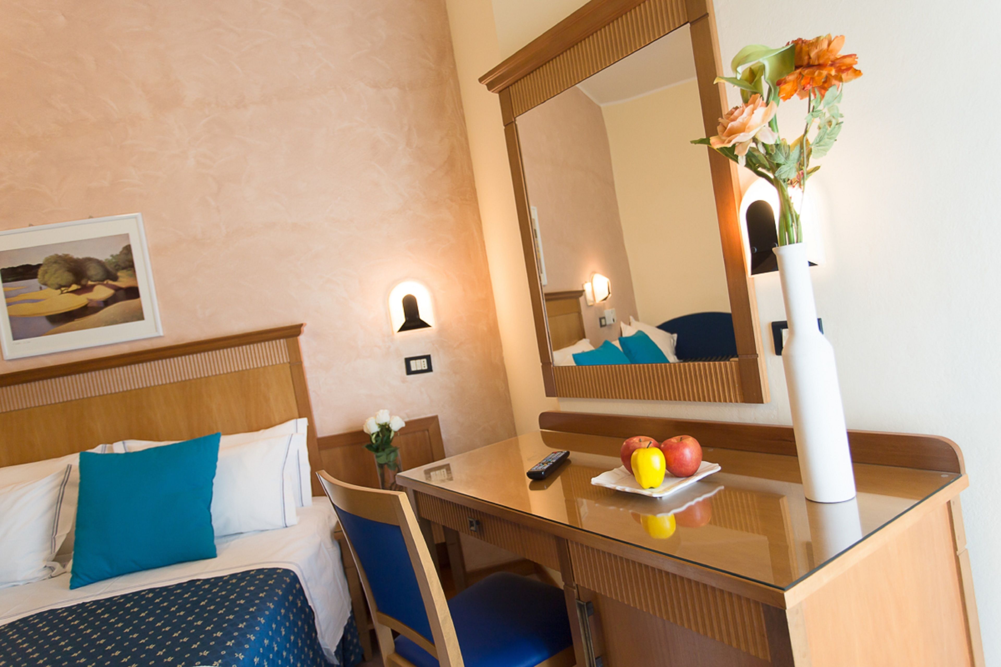 Une chambre d’hôtel avec un lit, un bureau, une chaise, un miroir, un vase avec des fleurs et une assiette de fruits.