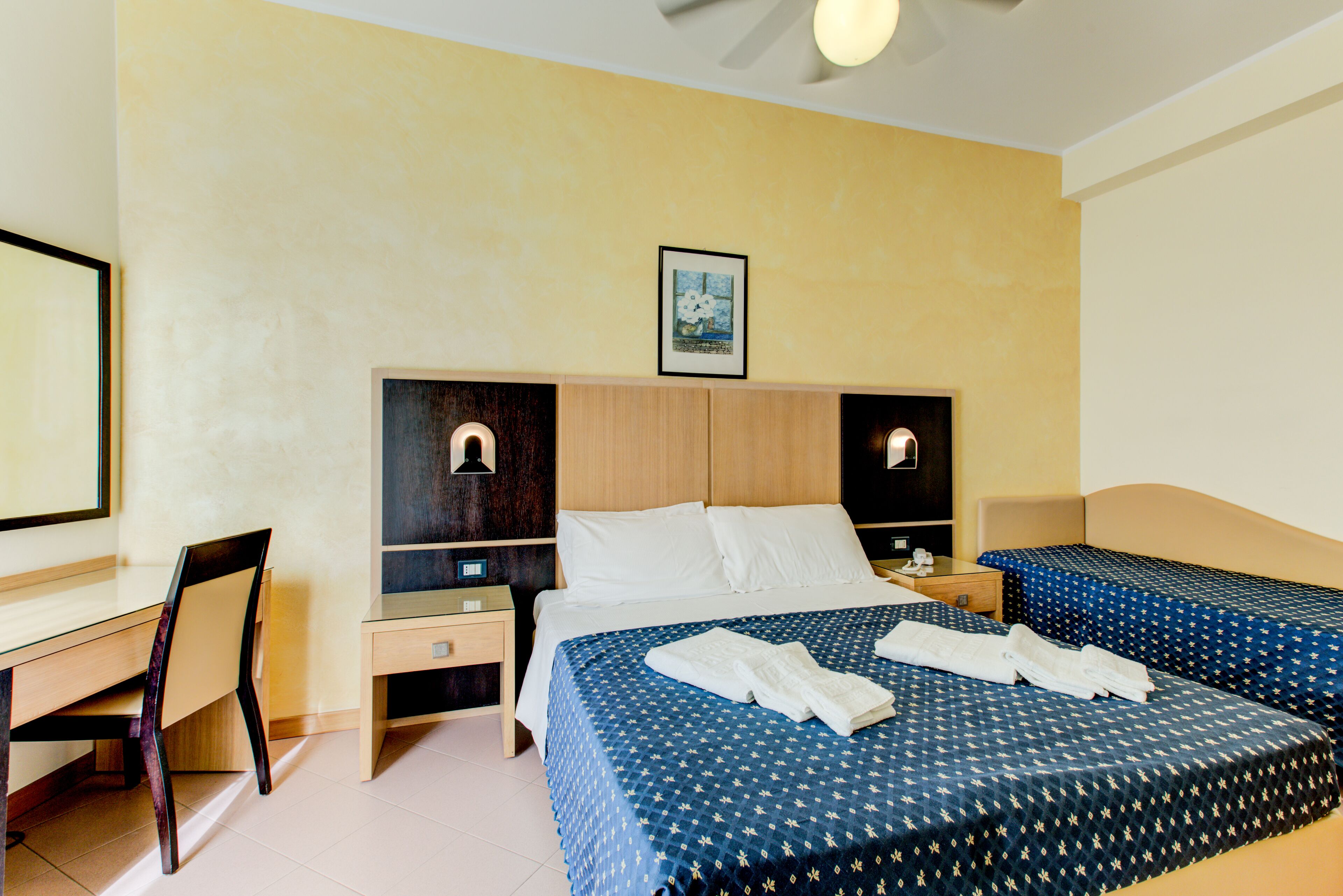 A hotel room with a bed, two bedside tables, a desk, a chair, and a ceiling fan.