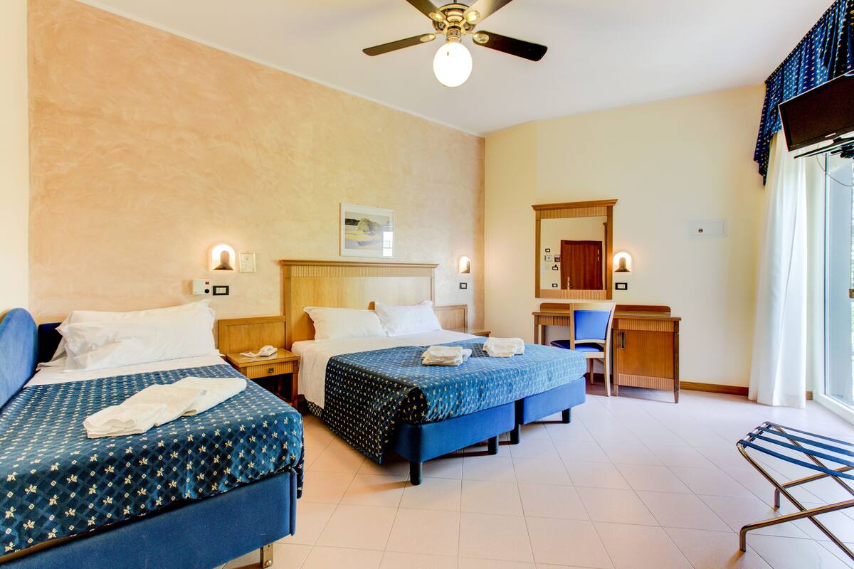 A hotel room with three beds, a desk, a chair, a ceiling fan, and a window with curtains.