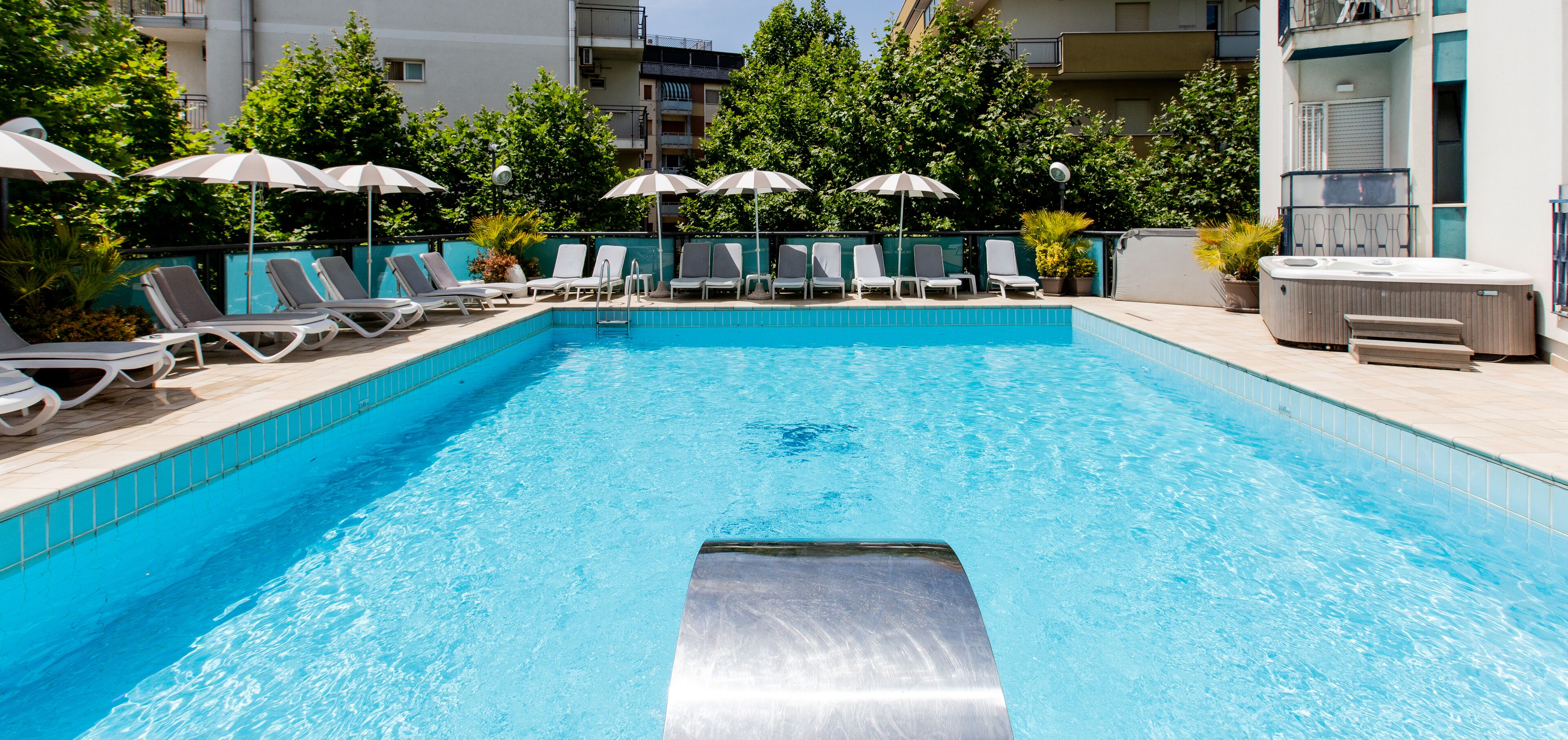 Une piscine entourée de verdure et de bâtiments, avec des chaises longues et des parasols.