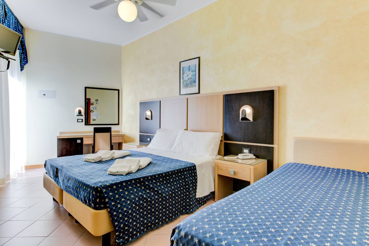 A hotel room with two beds, a desk, a chair, a ceiling fan, a bedside table with a lamp, and a framed picture on the wall.