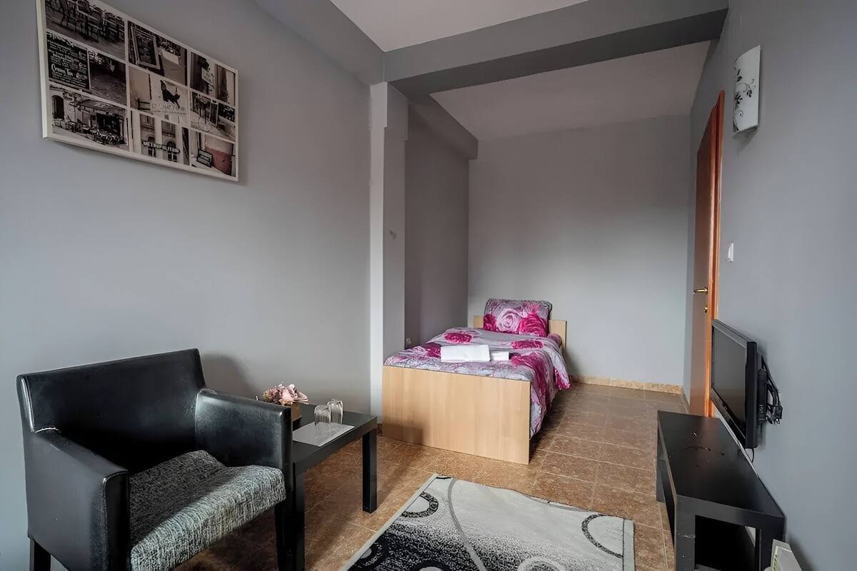 Une pièce de taille modeste comprenant un lit, une chaise, une petite table, un téléviseur et une œuvre d’art murale.