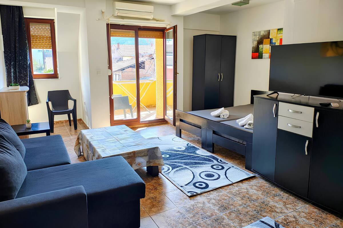 Un espace de vie compact comprenant un canapé, une petite table, un coin cuisine avec un téléviseur et une porte-fenêtre coulissante donnant sur un balcon.