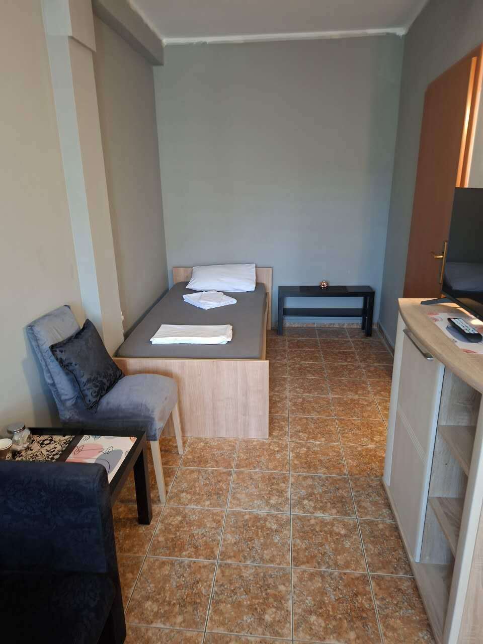 Una habitación pequeña con cama, una mesita, una silla, un banco y un televisor.