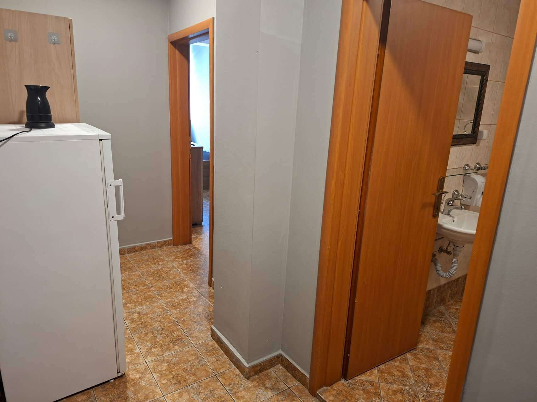 Ein schmaler Flur mit einem weißen Kühlschrank, einer Holztür und einem Badezimmer inklusive Waschbecken und Spiegel.