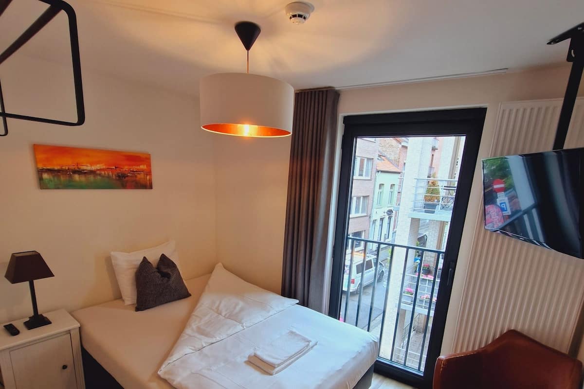 A hotel room with a bed, a nightstand, a TV, a chair, and a view of the outside.