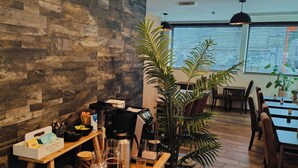 Un accogliente caffè con un bancone in legno, macchine da caffè e una varietà di bevande. Si vede un'ampia vetrata con persiane, un lampadario e una pianta in vaso.