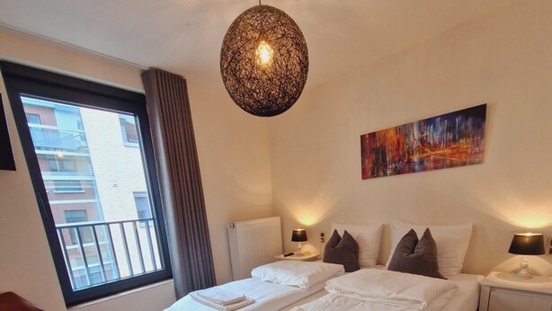 Una camera d'albergo moderna con un letto grande, una poltrona rossa, una finestra vista edifici e un lampadario.