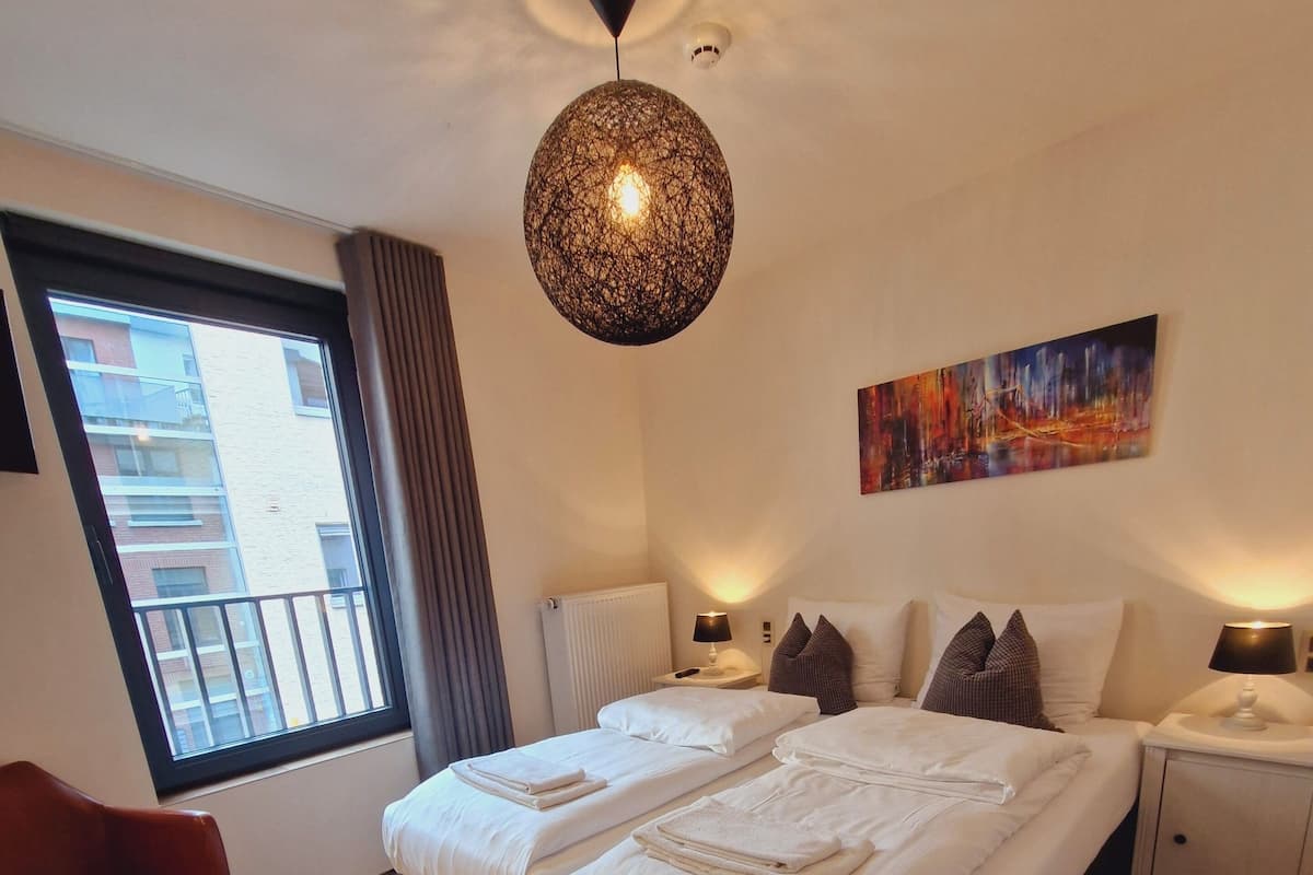 A modern hotel room with a large bed, a red chair, a window with a view of buildings, and a ceiling light fixture.