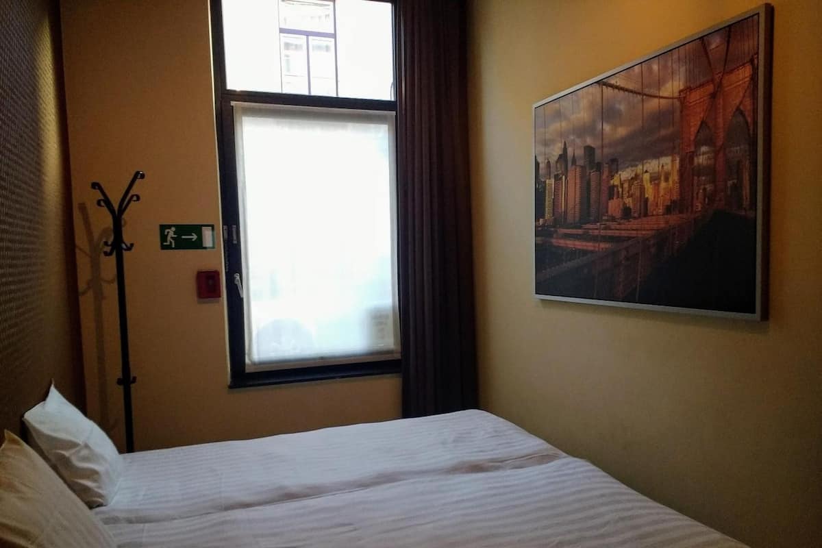 Una habitación de hotel con una cama, una ventana con cortinas, un perchero y un cuadro en la pared.