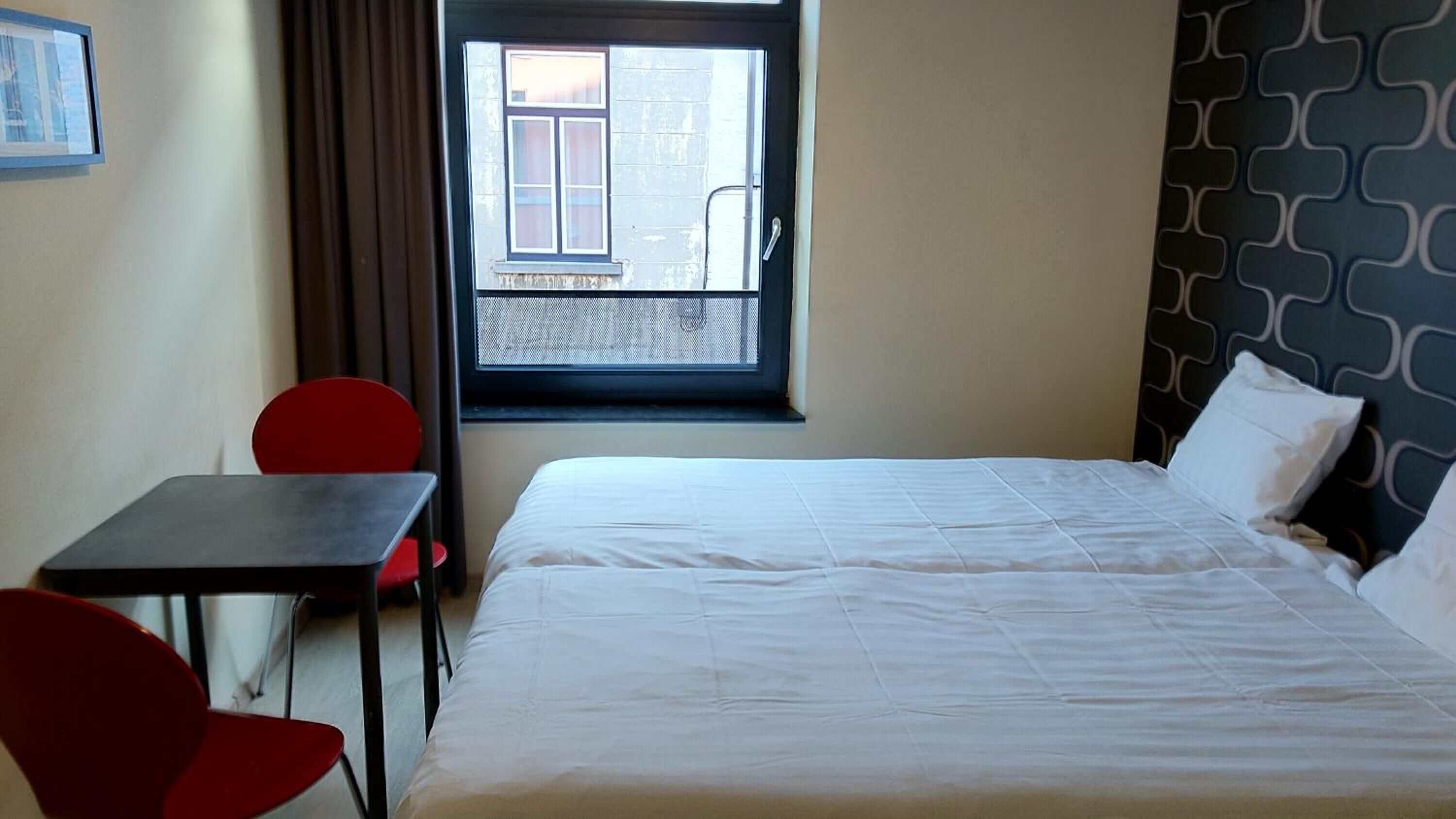 A hotel room with a bed, a small table, two red chairs, a window with a view of a balcony, and a patterned wall.