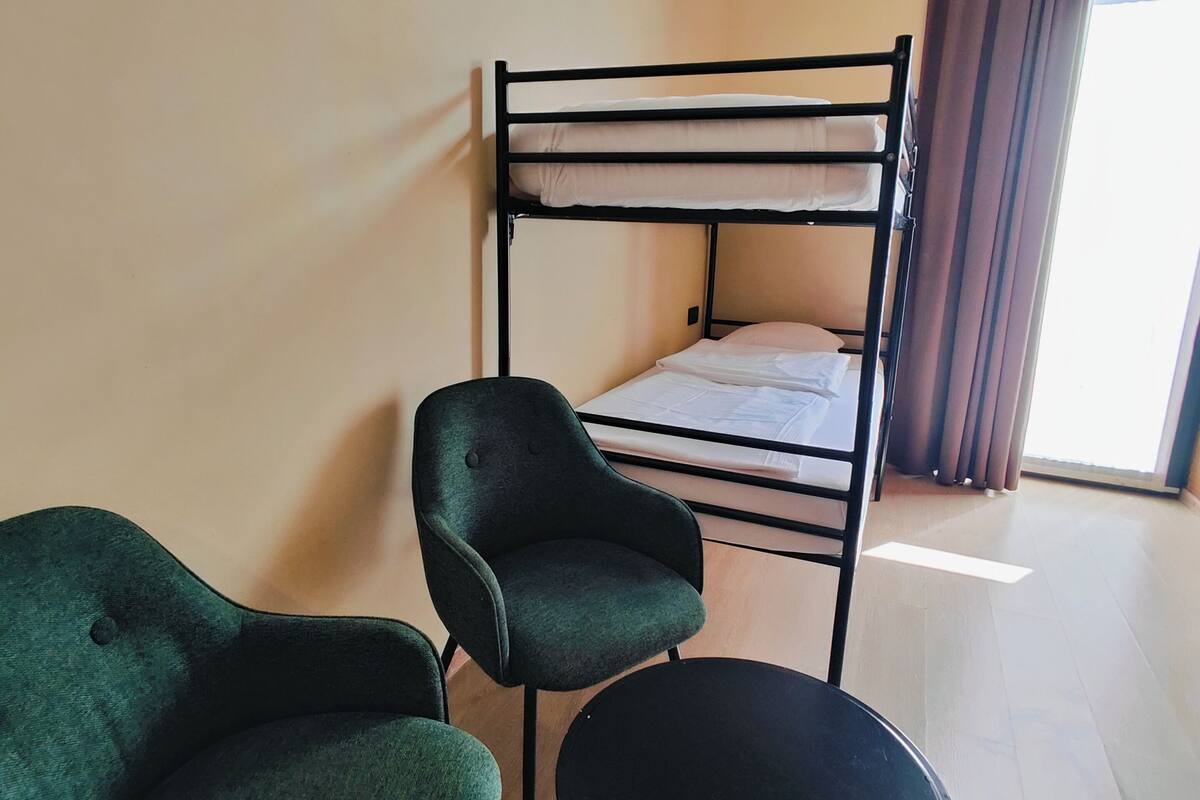 Una habitación con una cama litera, dos sillas verdes, una mesita negra y un ventanal con cortinas.
