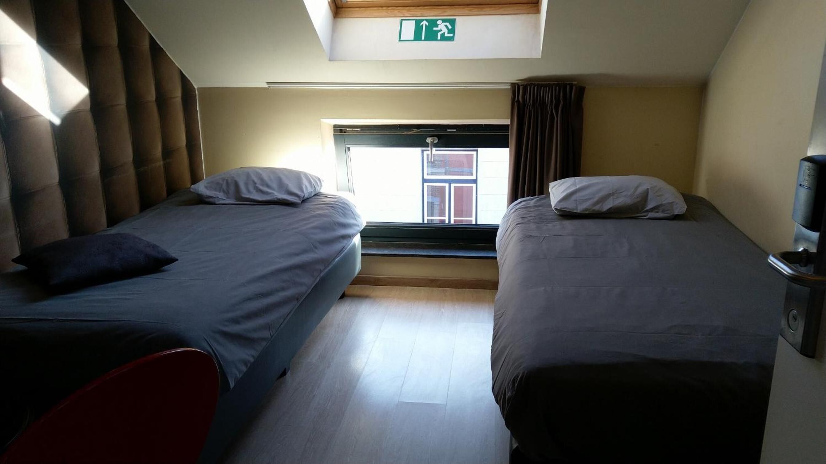 Een kamer met twee bedden, een raam met gordijnen, een deur en een pictogram voor een vluchtweg.