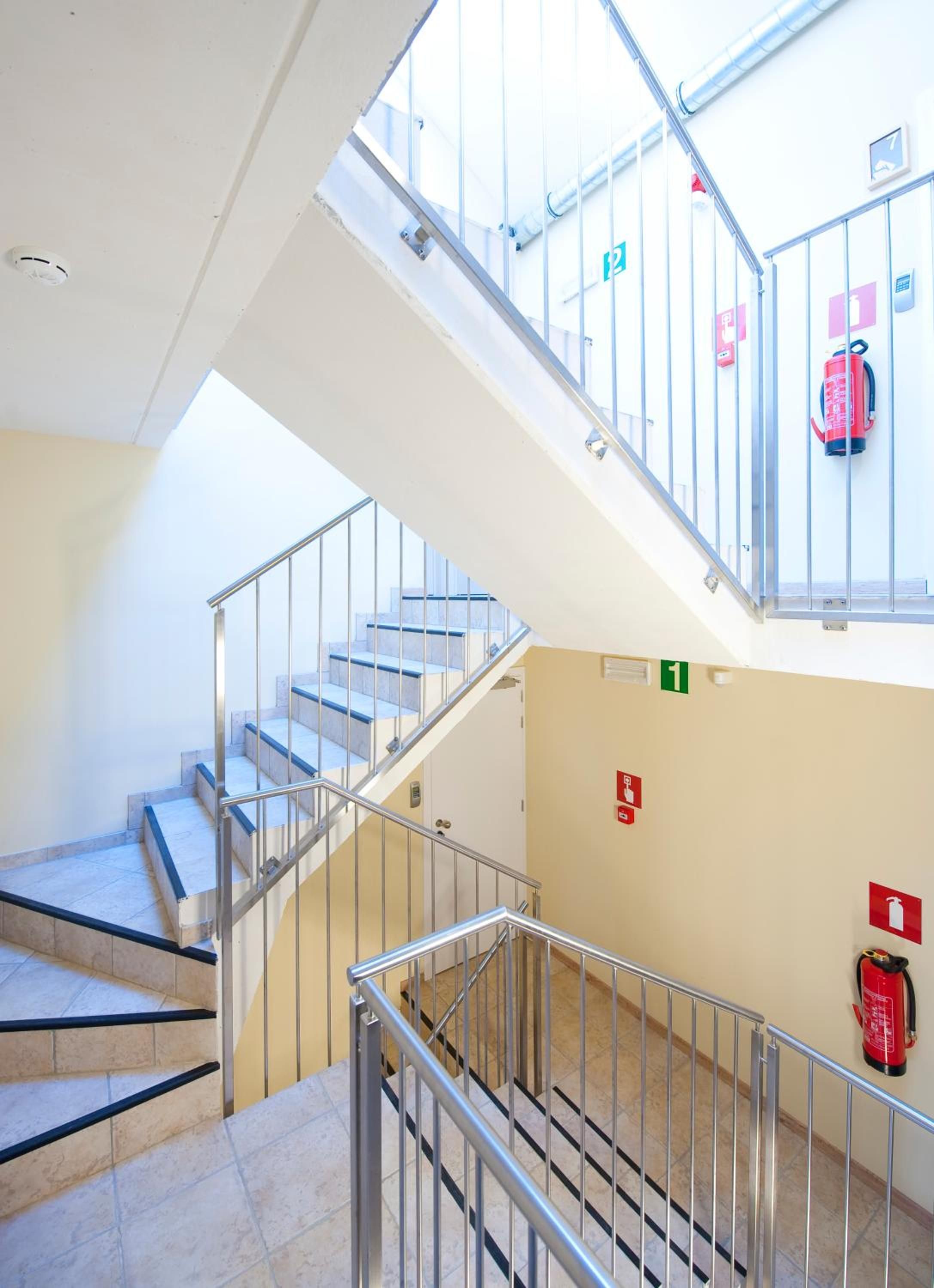 Un interior de varios niveles con escalera, barandillas metálicas y señales de salida de emergencia.