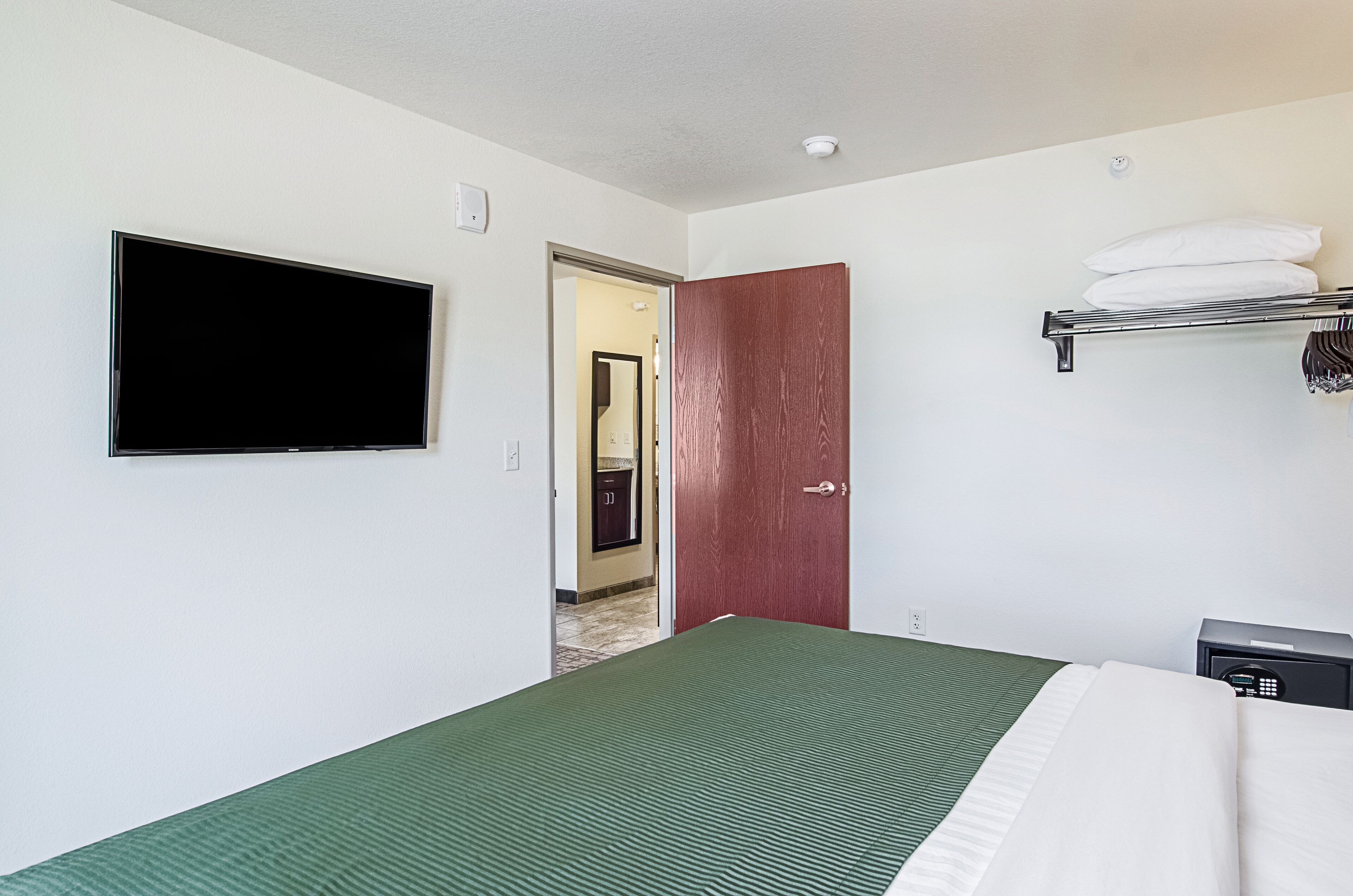 Quarto de hotel com cama, televisão na parede, porta e pequena mesa de cabeceira com abajur.