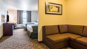 Een hotelkamer met een bruine bank, een bureau met een computer, een televisie, twee bedden en een ingelijst kunstwerk aan de muur.