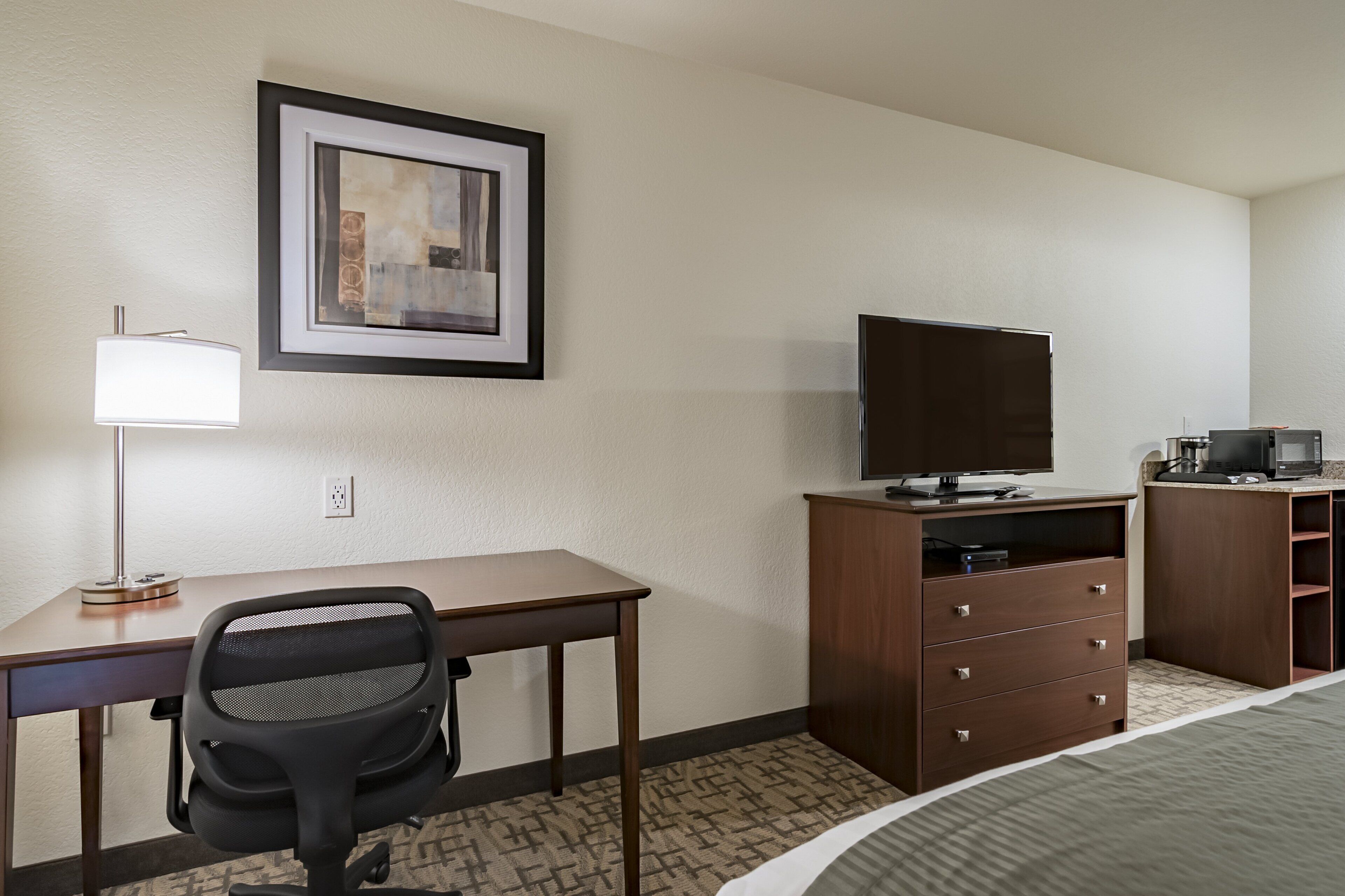 A hotel room with a desk, chair, television, and a dresser.
