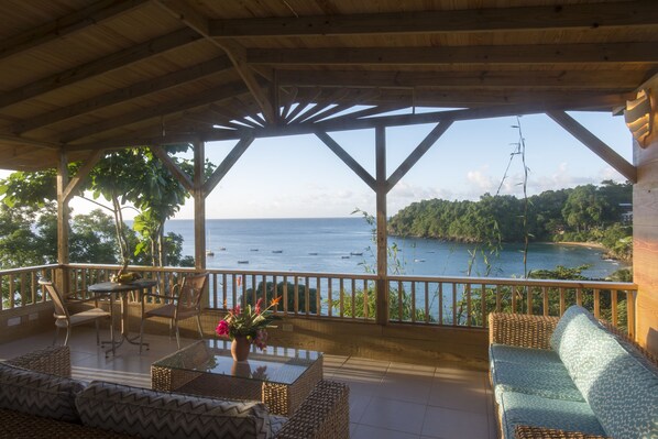 Un espace extérieur aménagé, abrité et doté de mobilier en osier, d’une table en verre, et offrant une vue sur la mer et la verdure.